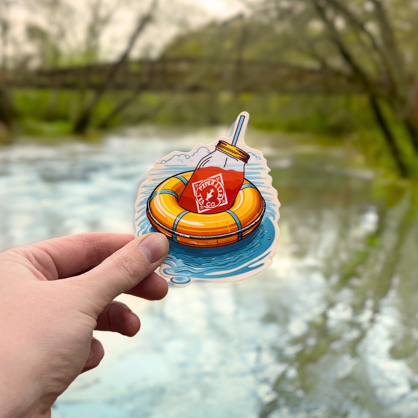 A hand holds the "Life Preserver with Quart Jar Sticker" by Piper & Leaf Tea Co., depicting an orange inflatable ring and iced tea floating on water, set against a serene river backdrop, exuding summer bliss.