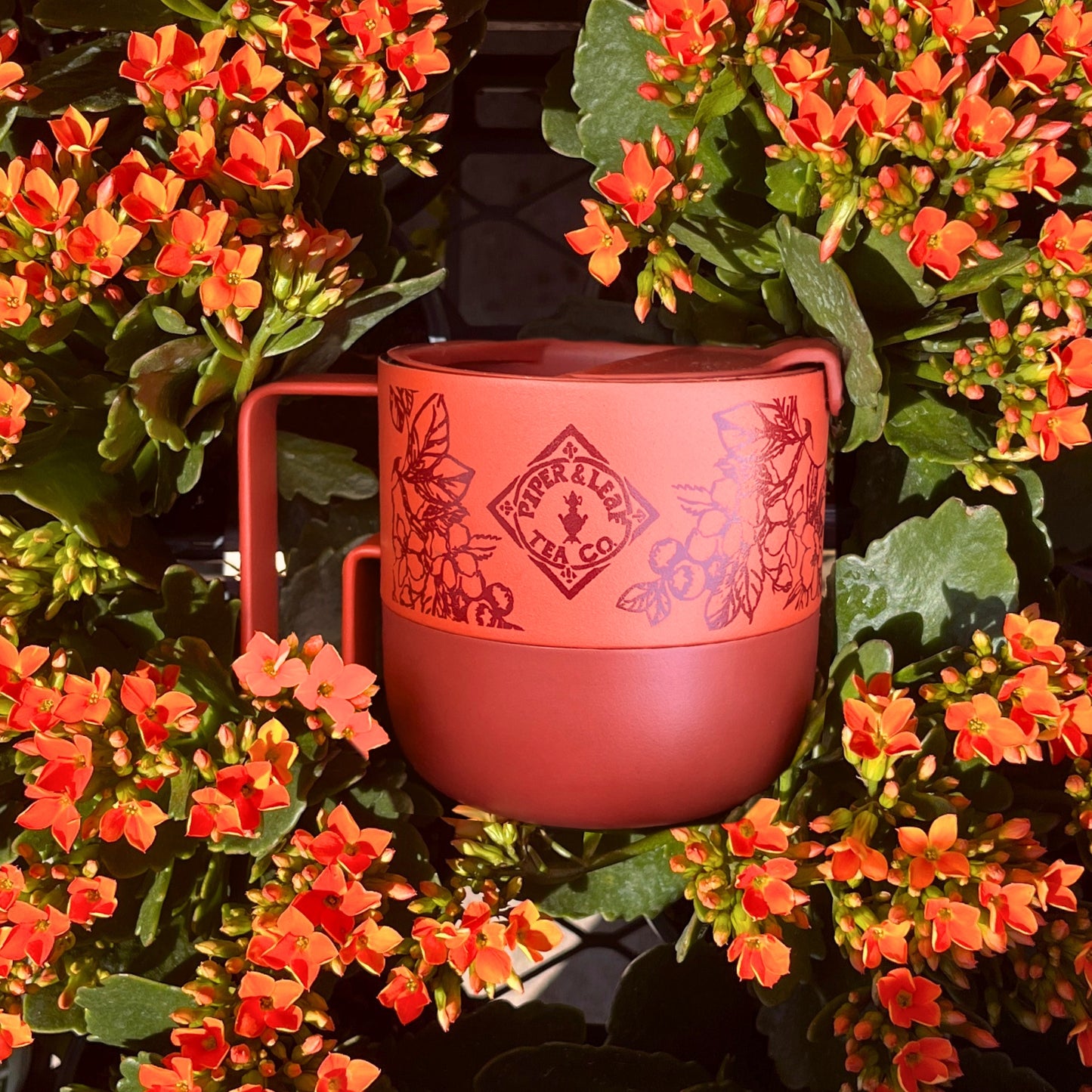 The P&L 14oz Rise Mug from Piper & Leaf Tea Co. by Klean Kanteen, adorned with floral patterns and a logo, is perfect for tea and sits among vibrant orange flowers and green leaves.
