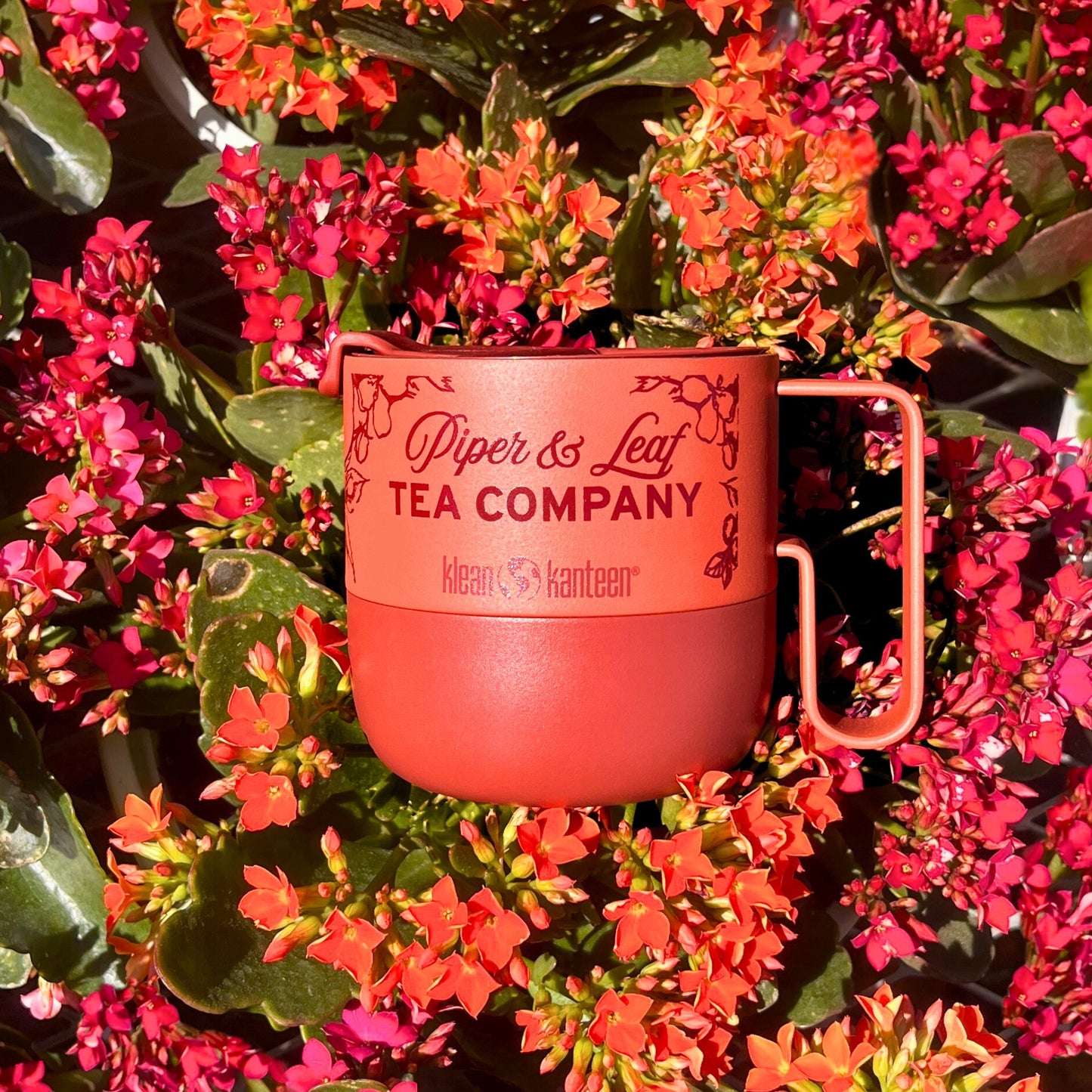 The P&L 14oz Rise Mug from Klean Kanteen, by Piper & Leaf Tea Co., is a red metal mug made from recycled stainless steel, nestled among pink and red flowers—perfect for tea lovers who value nature's beauty and eco-friendly choices.