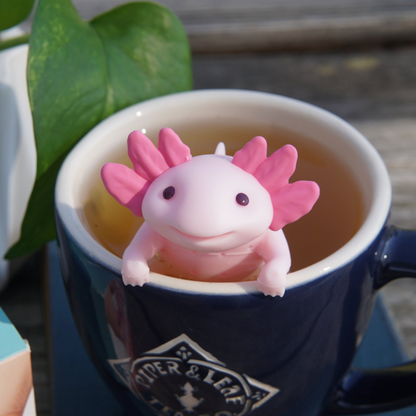 A Piper & Leaf Tea Co. Relaxolotl-Tea Infuser, shaped like a charming pink axolotl, sits on the edge of a navy blue cup of tea, amidst lush green leaves.