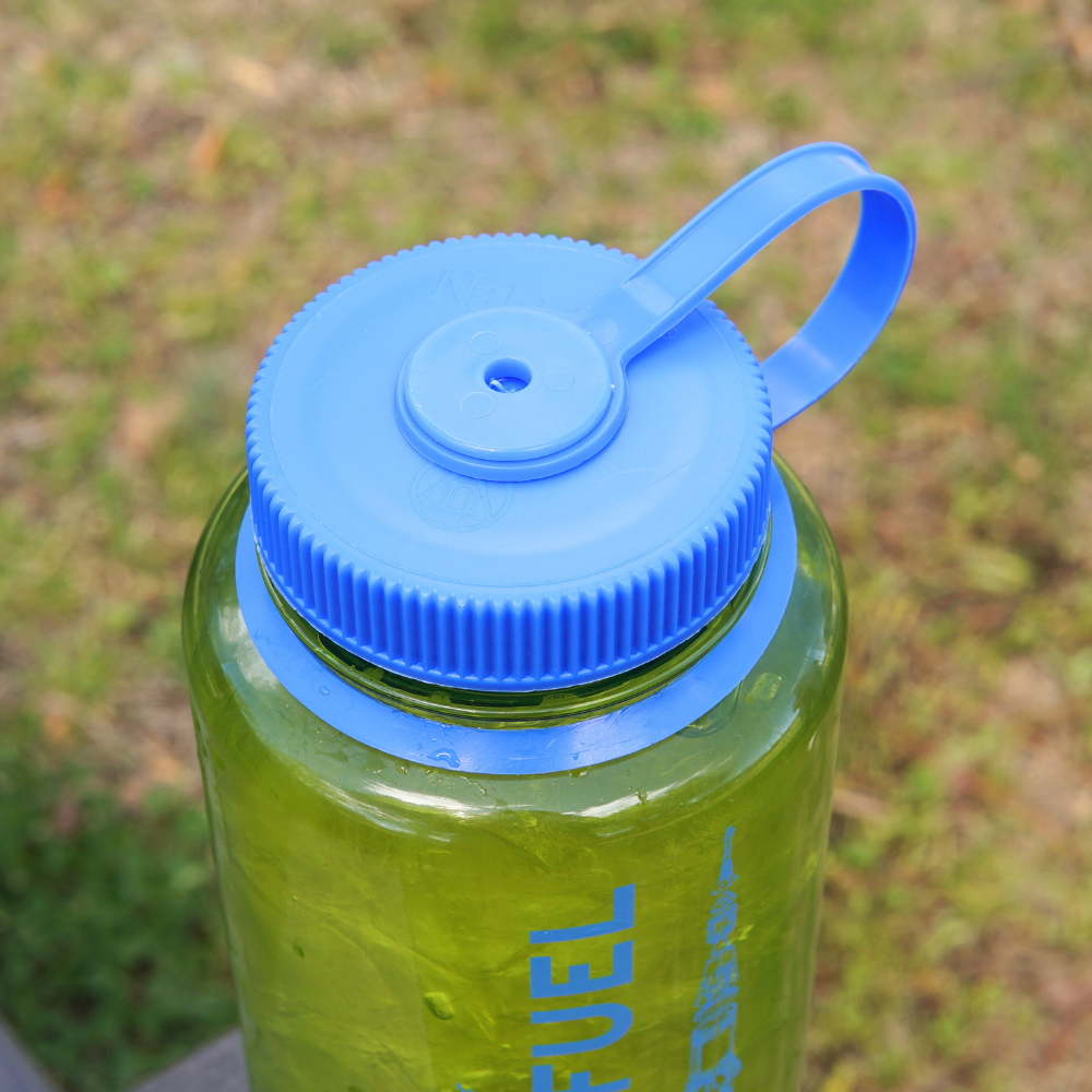Blue lid on a green Piper & Leaf Nalgene Water (Tea) Bottle- Rocket Fuel Edition with a loop handle, set outdoors.