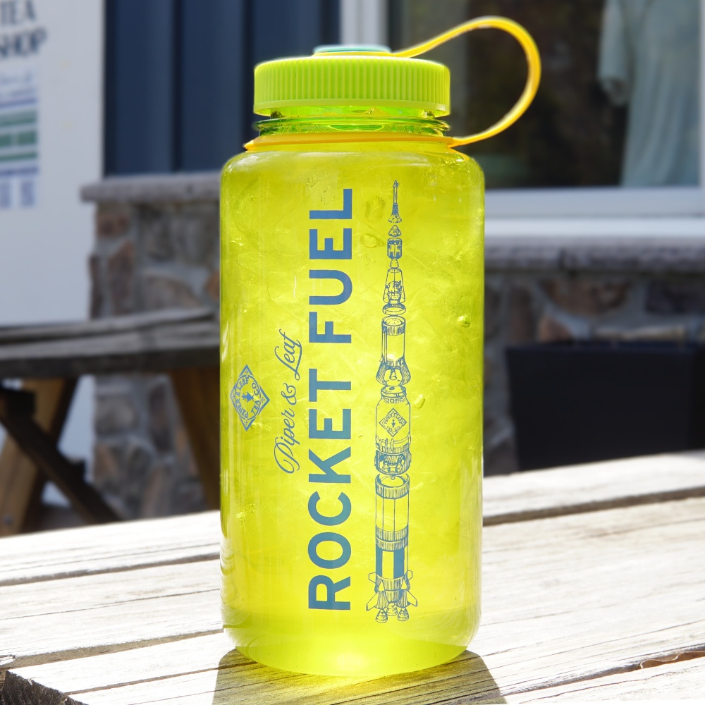 A translucent yellow Piper & Leaf Nalgene Water (Tea) Bottle- Rocket Fuel Edition, featuring an illustration of a rocket on it, sitting on a wooden surface outdoor.