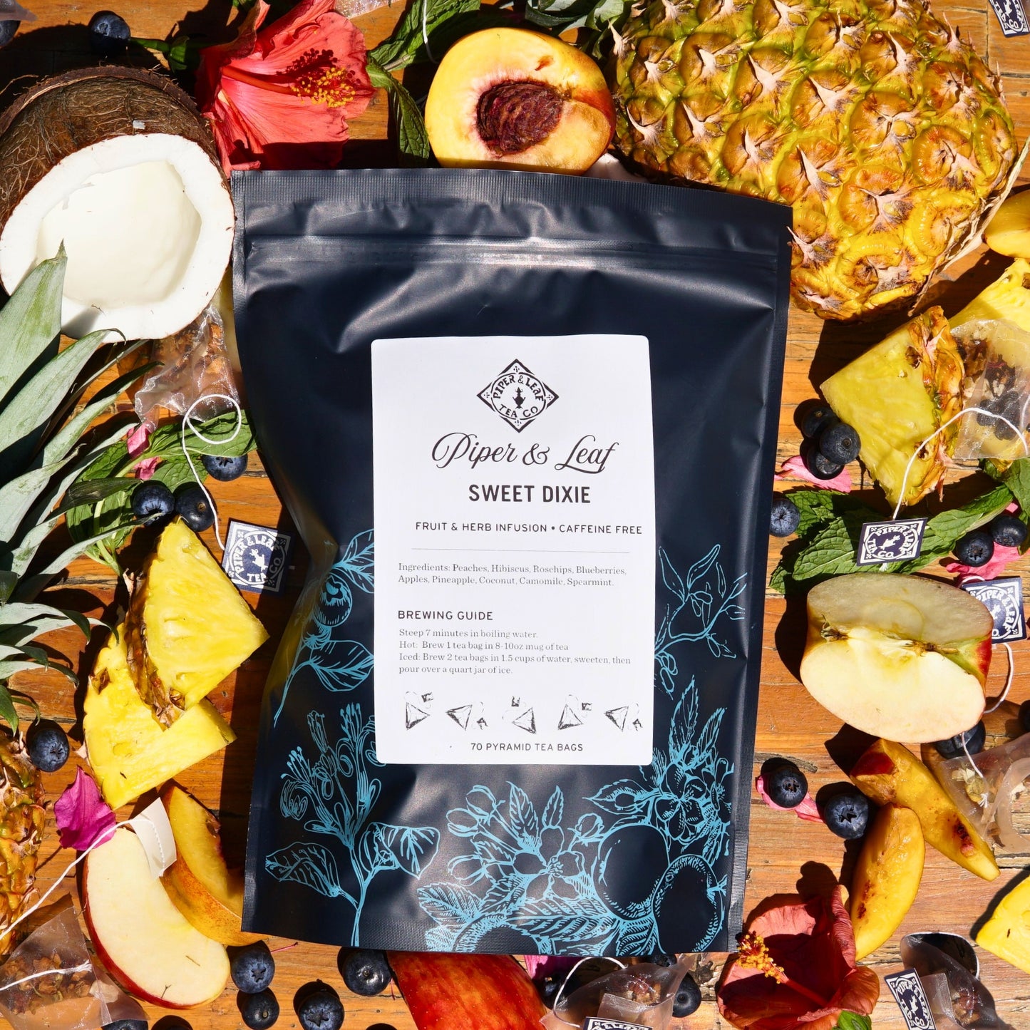 A black bag of Sweet Dixie Bulk Sachets from Piper & Leaf Tea Co. sits on a wooden surface, surrounded by vibrant fruits like pineapples, peaches, blueberries, and coconut halves—a perfect caffeine-free summer brew.