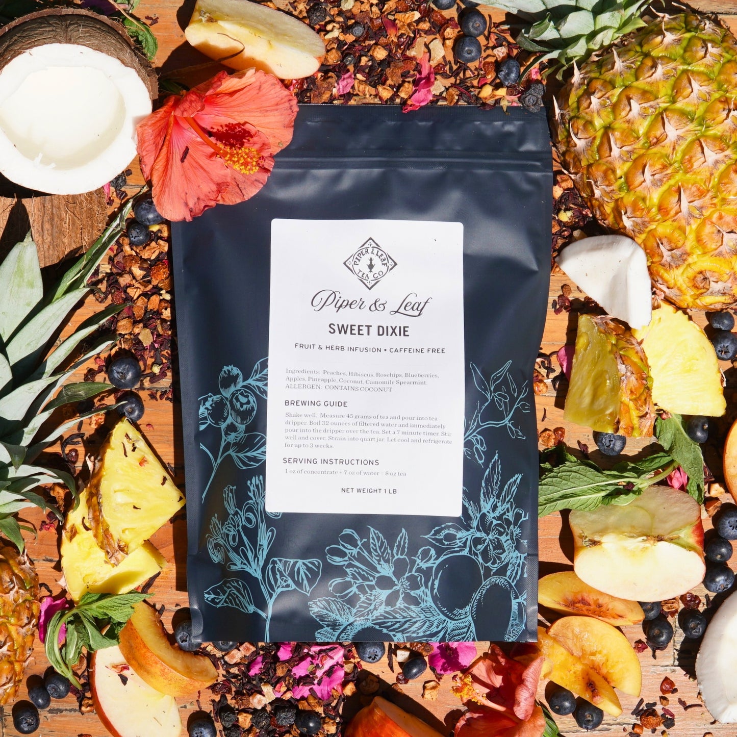 A bag of Piper & Leaf Tea Co.'s Sweet Dixie Pound Bag—190 servings—sits enveloped by a vibrant fruit medley with loose leaf tea, berries, pineapple, apples, and hibiscus on a wooden surface—a perfect summertime brew.