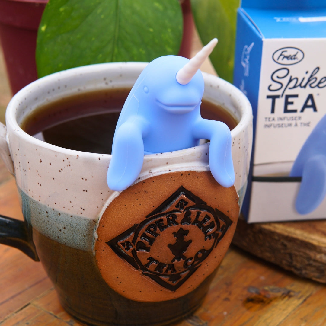 A delightful blue narwhal-shaped tea strainer from Piper & Leaf Tea Co., named the Spiked Tea Infuser, graces your mug, steeping your tea perfectly. Nearby, a touch of greenery and the infuser box enhance the serene atmosphere.