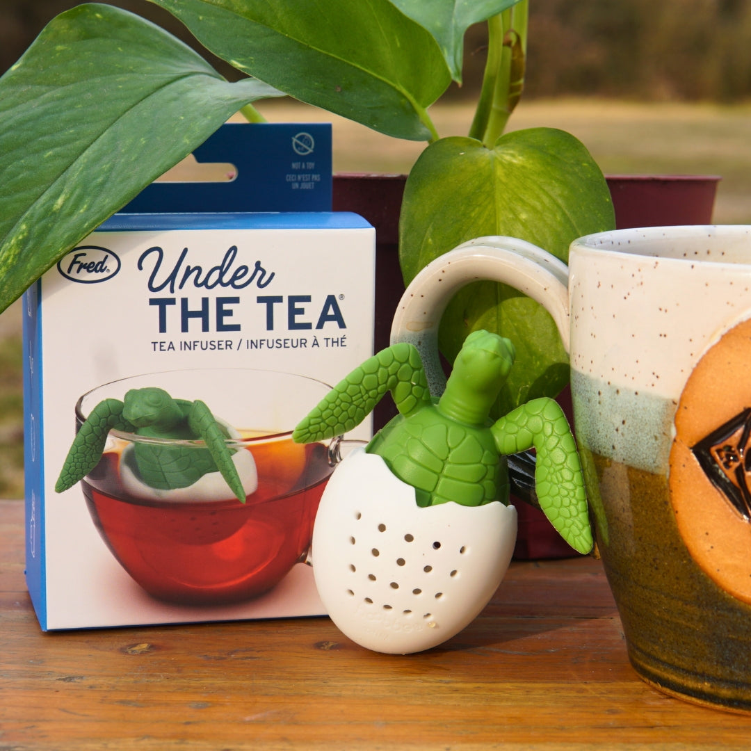 The Under the Tea Infuser by Piper & Leaf Tea Co., shaped like a turtle, sits on a wooden surface with its packaging next to a ceramic mug. Reusable metal straws and a potted plant add to the scene.