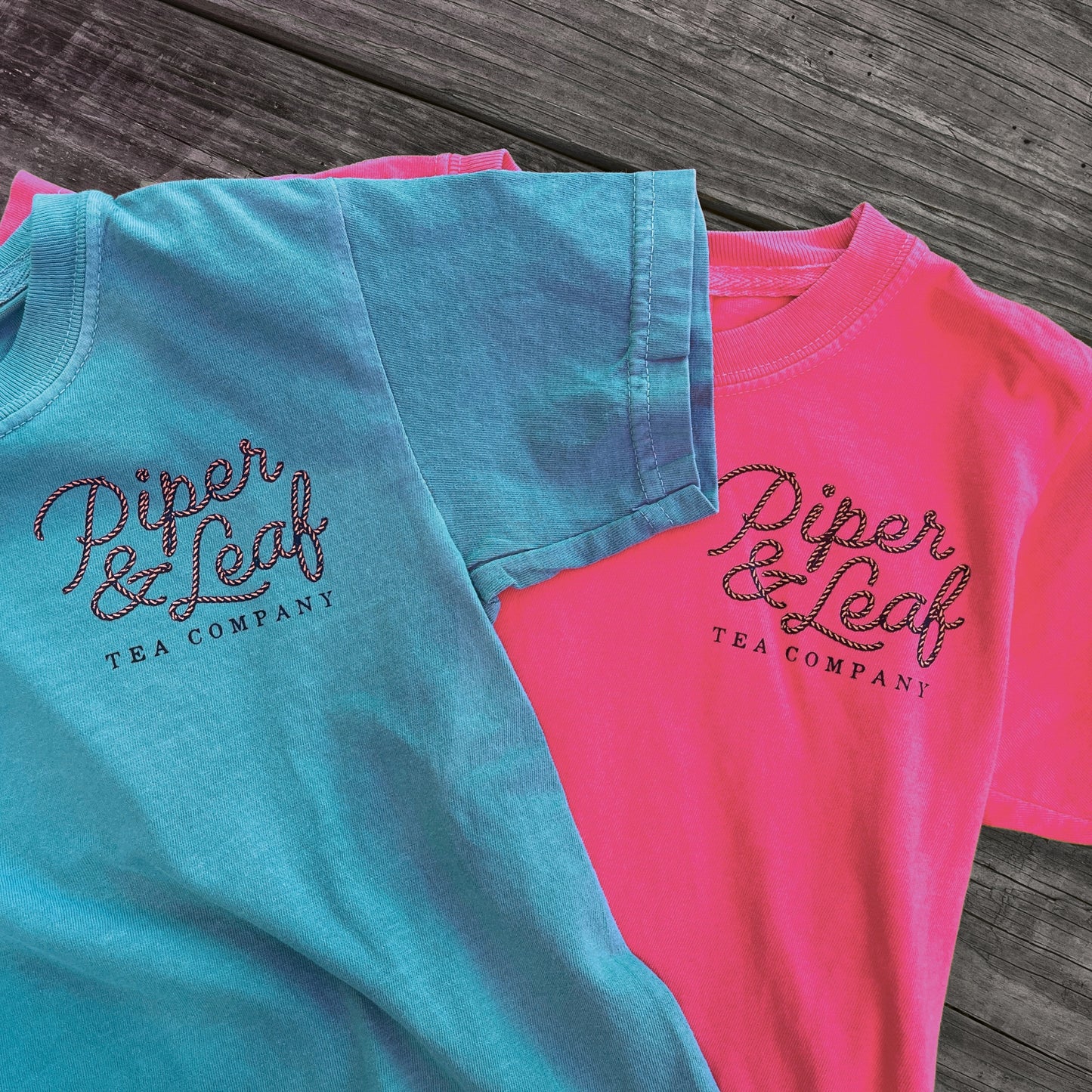 Two Life Preserver T-Shirts by Piper & Leaf Tea Co.—one teal, one pink—are laid flat on a wooden surface, ready to wear for sipping iced tea on a summer day.