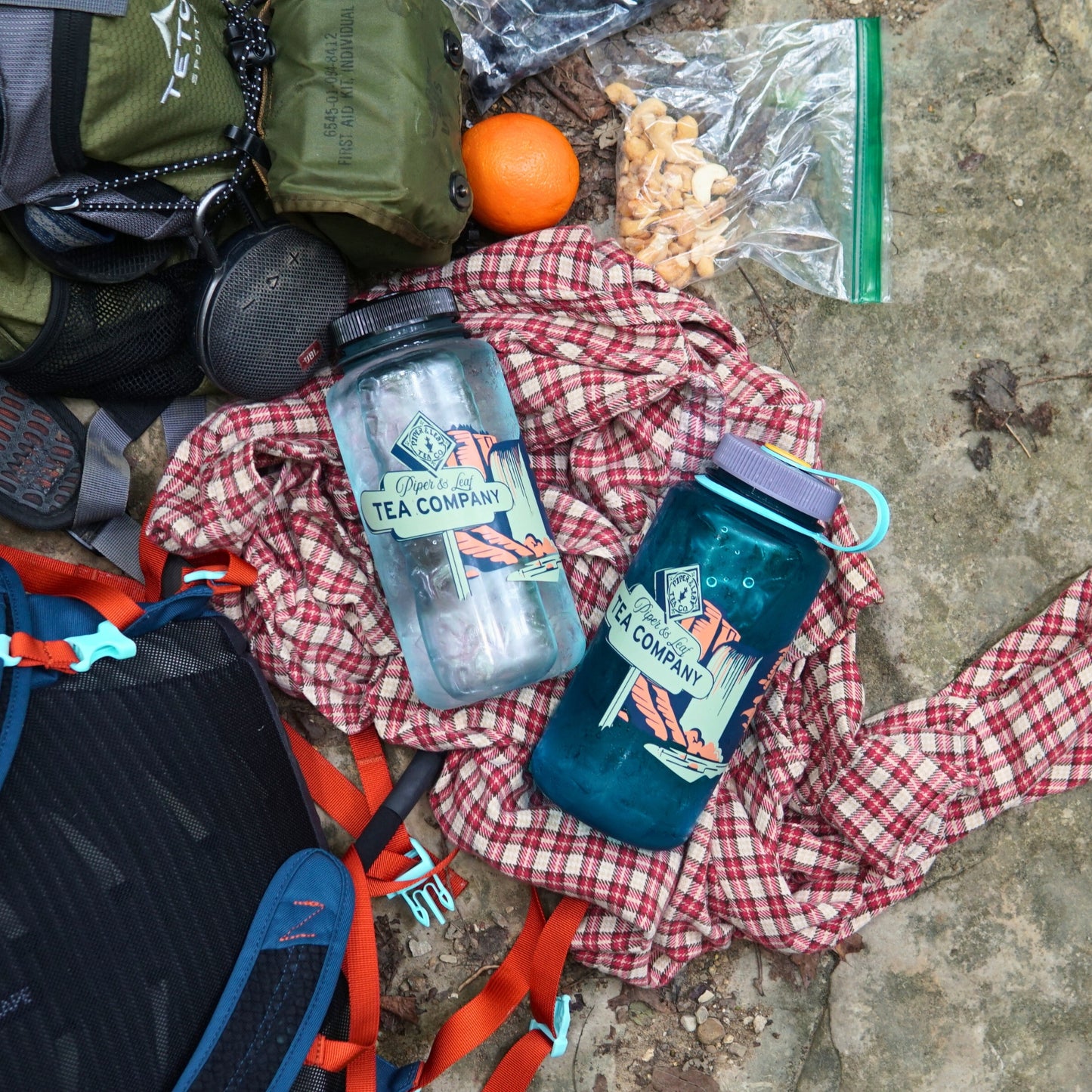 Two Piper & Leaf Tea Co. "Tea, Trails and Tall Tales" Nalgene bottles sit on a plaid shirt, accompanied by a backpack, hiking gear, tea, nuts, and an orange—all ready for your next rocky adventure.