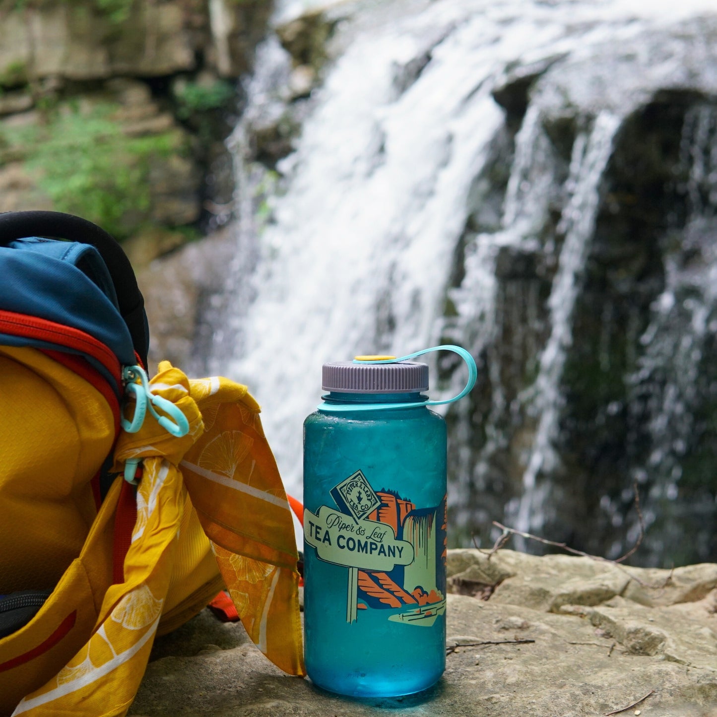 A Tea, Trails and Tall Tales Nalgene by Piper & Leaf Tea Co. rests on a rock by a forest waterfall, ready to fuel your next adventure.
