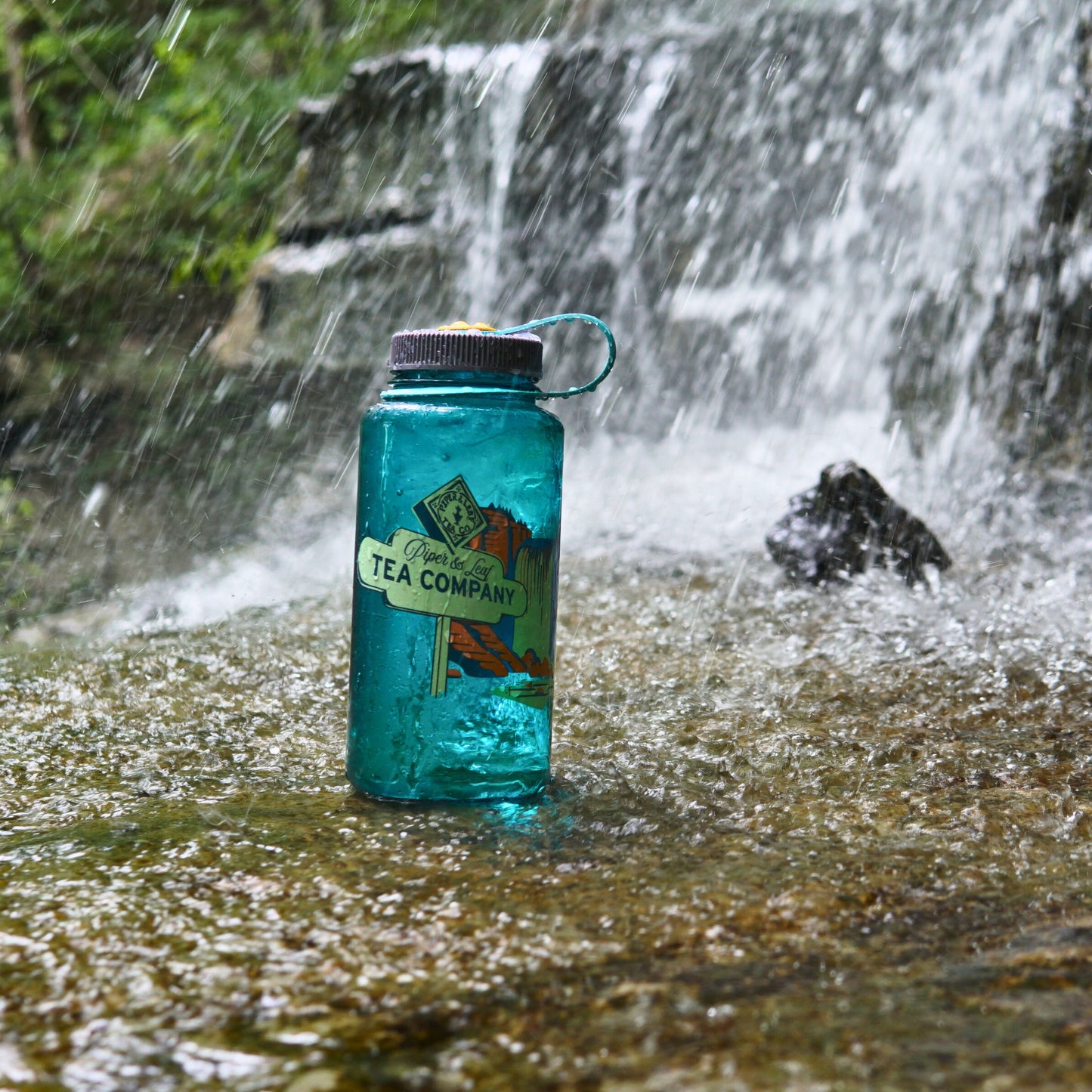 A Piper & Leaf Tea Co. "Tea, Trails and Tall Tales" Nalgene sits on a wet rock by a small waterfall, ready to join you on your next outdoor adventure.