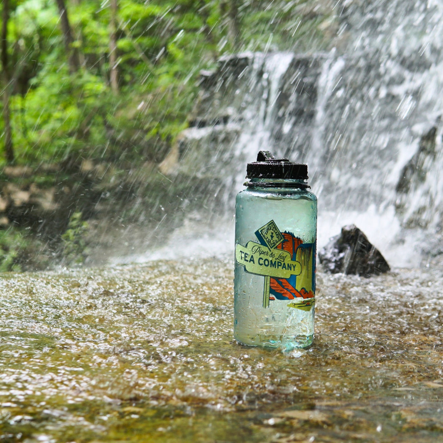 The Piper & Leaf Tea Co. "Tea, Trails and Tall Tales" Nalgene bottle sits on a wet rock by a waterfall, vibrant label shining amid lush greenery—perfect for your next outdoor adventure.