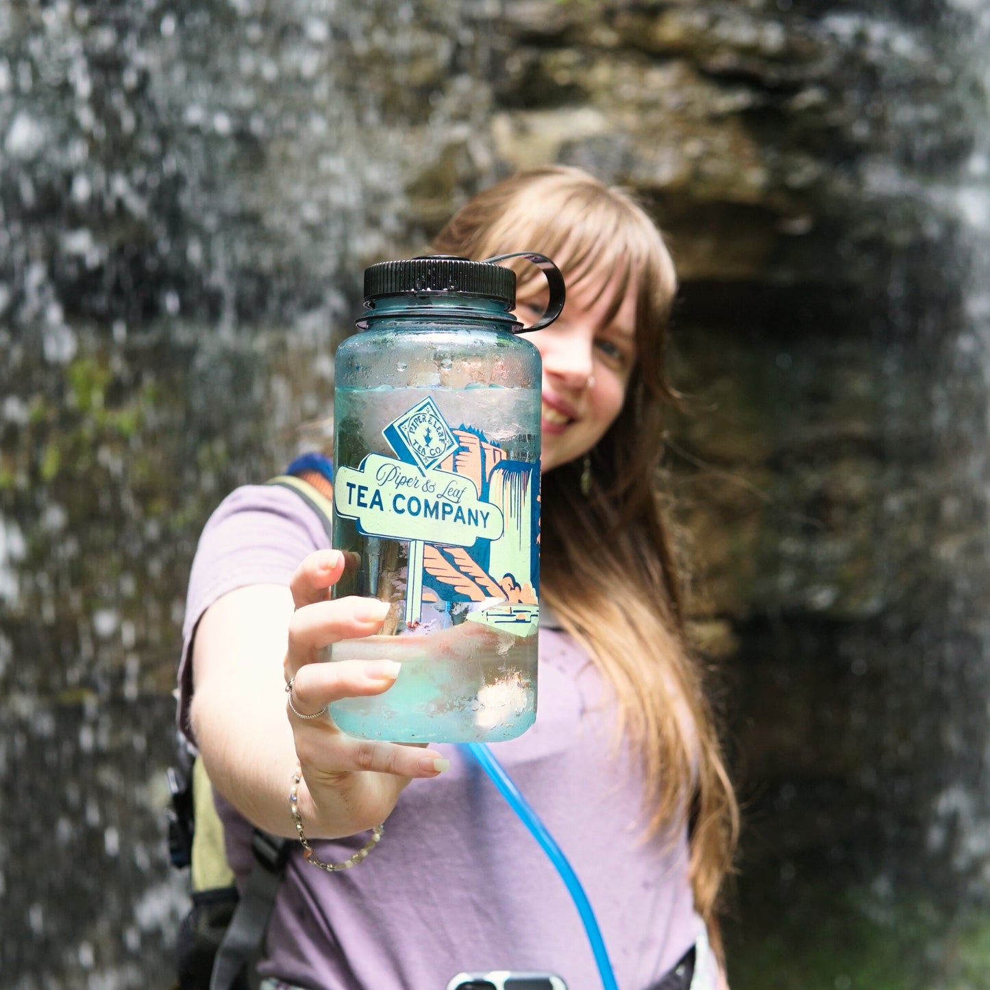 A woman with long hair stands by a waterfall, holding a clear "Tea, Trails and Tall Tales Nalgene" bottle from Piper & Leaf Tea Co. toward the camera, capturing an adventurous outdoor spirit.