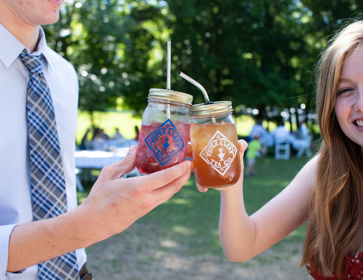 Two people clinking pint-sized Signature Mason Jars filled with iced tea and straws outdoors. The jars proudly display a logo for Piper & Leaf Tea Co., highlighting their dedication to sustainable farmers.