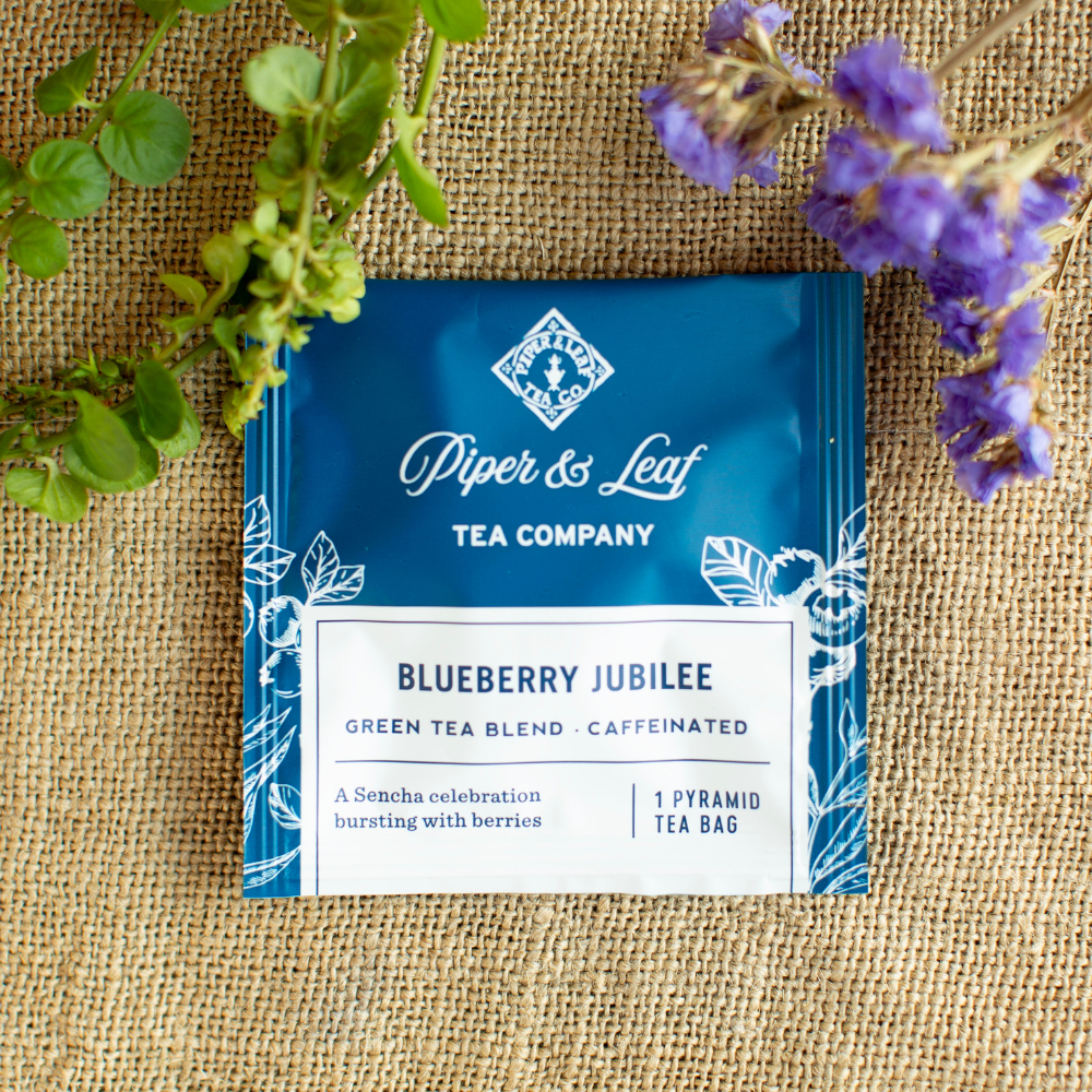 A Piper & Leaf Tea Co. Blueberry Jubilee box of 12 individually wrapped tea bag sachets rests on burlap amid green and purple foliage, showcasing its unique blueberry tea blend made with Sencha green tea.