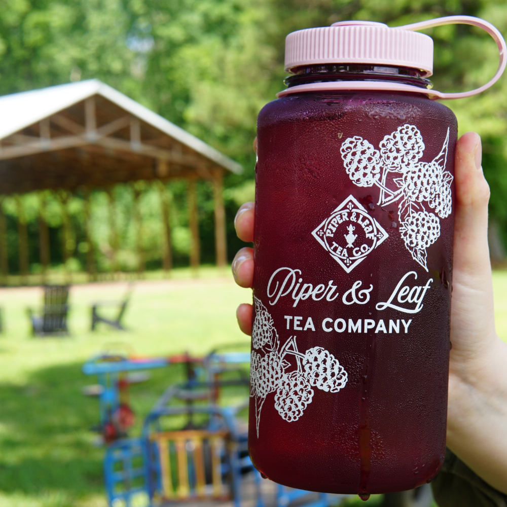 A hand holding a large translucent 32oz Wide Mouth Piper & Leaf Nalgene Water (Tea) Bottle - Into The Briar Patch, filled with a dark beverage, labeled "Piper & Leaf Tea Co." The background shows outdoor seating and a covered structure with trees in the distance, creating the perfect setting to enjoy your Briar Patch Brew.