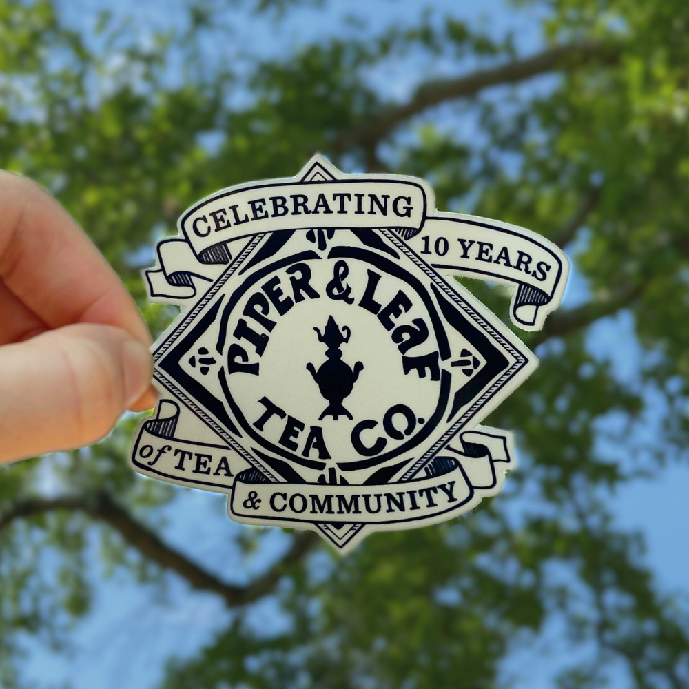 Hand holding a P&L 10 Year Anniversary Sticker with the text "Celebrating 10 Years of Tea & Community, Piper & Leaf Tea Co." against a background of green tree leaves and blue sky, marking this special anniversary.
