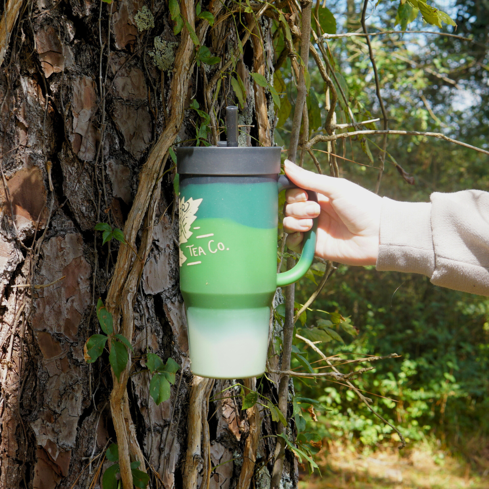 A hand holds a Piper & Leaf Tea Co. Limited Edition Autumn Leaf P&L X Silipint 32oz Tumbler with Handle, designed for seasonal sipping and featuring a sleek black lid and straw. This exclusive tumbler stands out against a tree draped in vines.