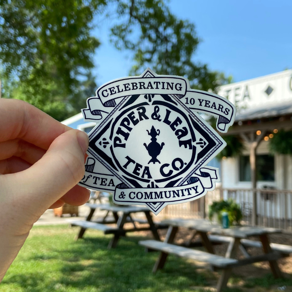 A hand holds a P&L 10 Year Anniversary Sticker by Piper & Leaf Tea Co., celebrating tea and community, with an outdoor seating area and trees in the background.