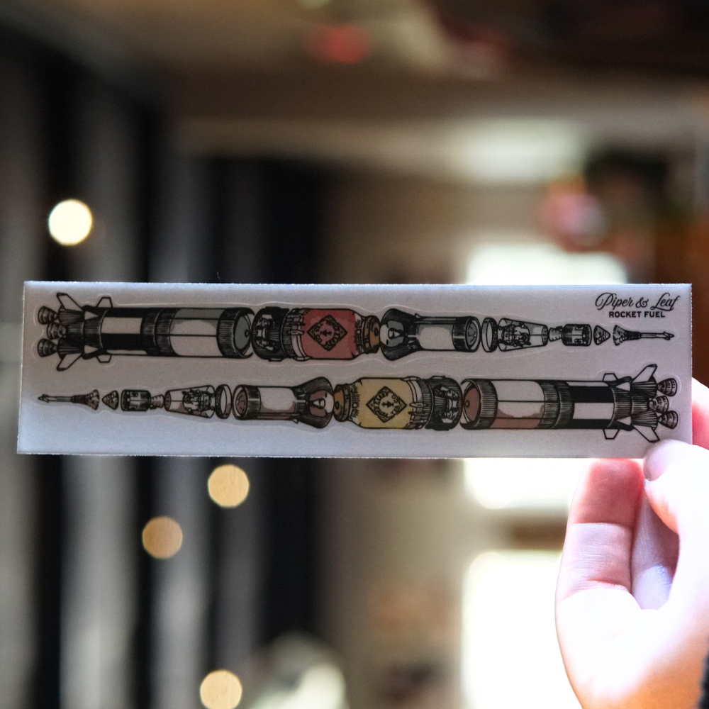 A person holding up a piece of paper with illustrations of two fancy art-deco-style pens along with the words "P&L Saturn V Rocket Fuel 6" Sticker Two Pack" and "Piper & Leaf Tea Co" written on it.