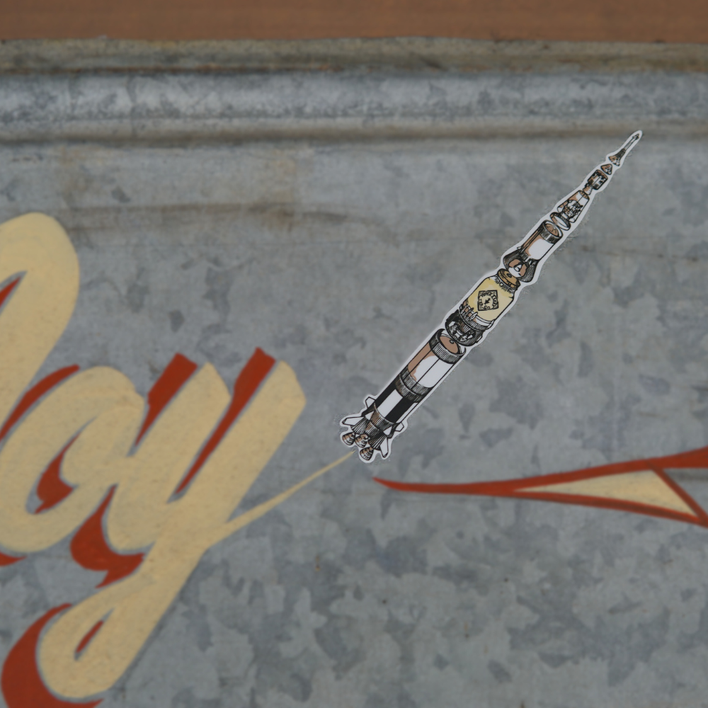 Silver and black pen with intricate designs placed on a wooden surface with the word "play" partially visible in the background, near a Piper & Leaf Tea Co P&L Saturn V Rocket Fuel 6" Sticker Two Pack.