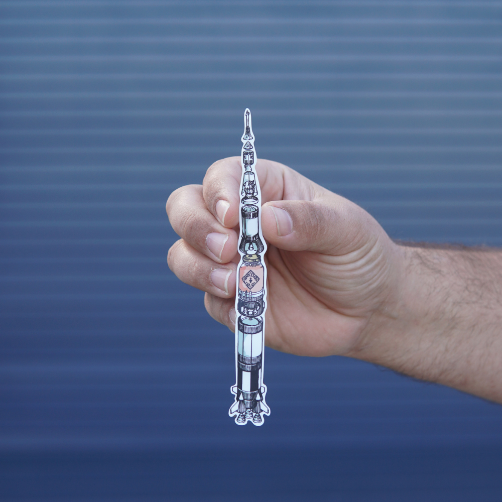 A hand holding a cutout illustration of a P&L Saturn V Rocket Fuel 6" Sticker Two Pack from Piper & Leaf Tea Co in front of a blue corrugated metal background.