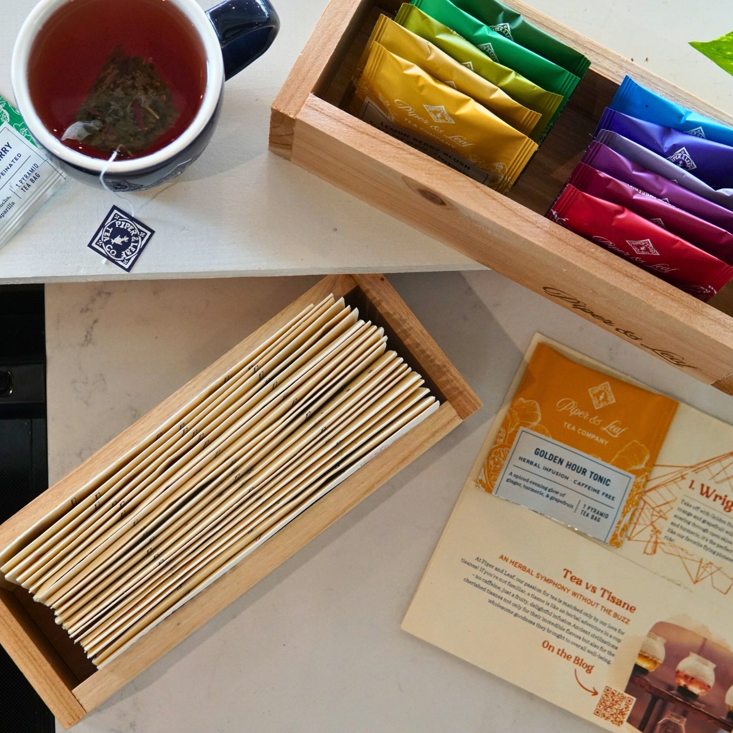 A cozy tea setup includes a steaming mug alongside a Piper & Leaf Tea Co. Wooden Steep Box packed with assorted tea bags, another box with wooden stir sticks, and an organized tea menu displayed neatly on the table to guide your choices.