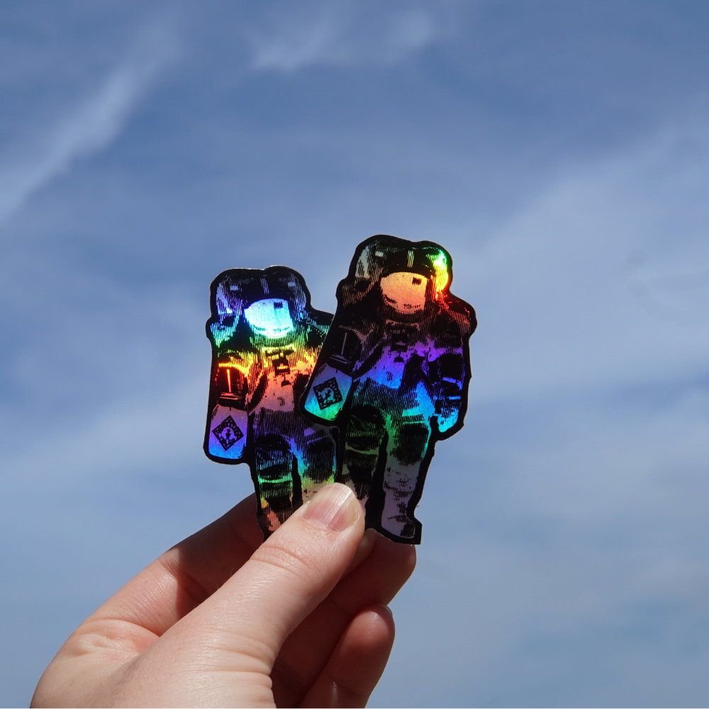 A hand holding two colorful, holographic spaceman keychains from the P&L Spaceman Holographic Sticker collection by Piper and Leaf Tea Co. against a clear blue sky.