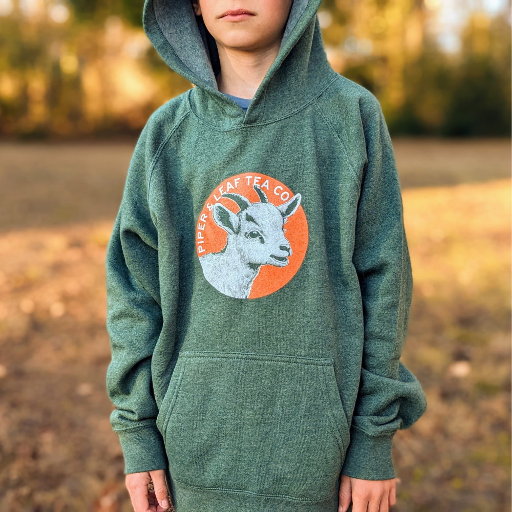A child wearing a green "Piper & Leaf Tea Co. Kids Being Kids- Children's Hoodie" with a deer logo on it stands outdoors.