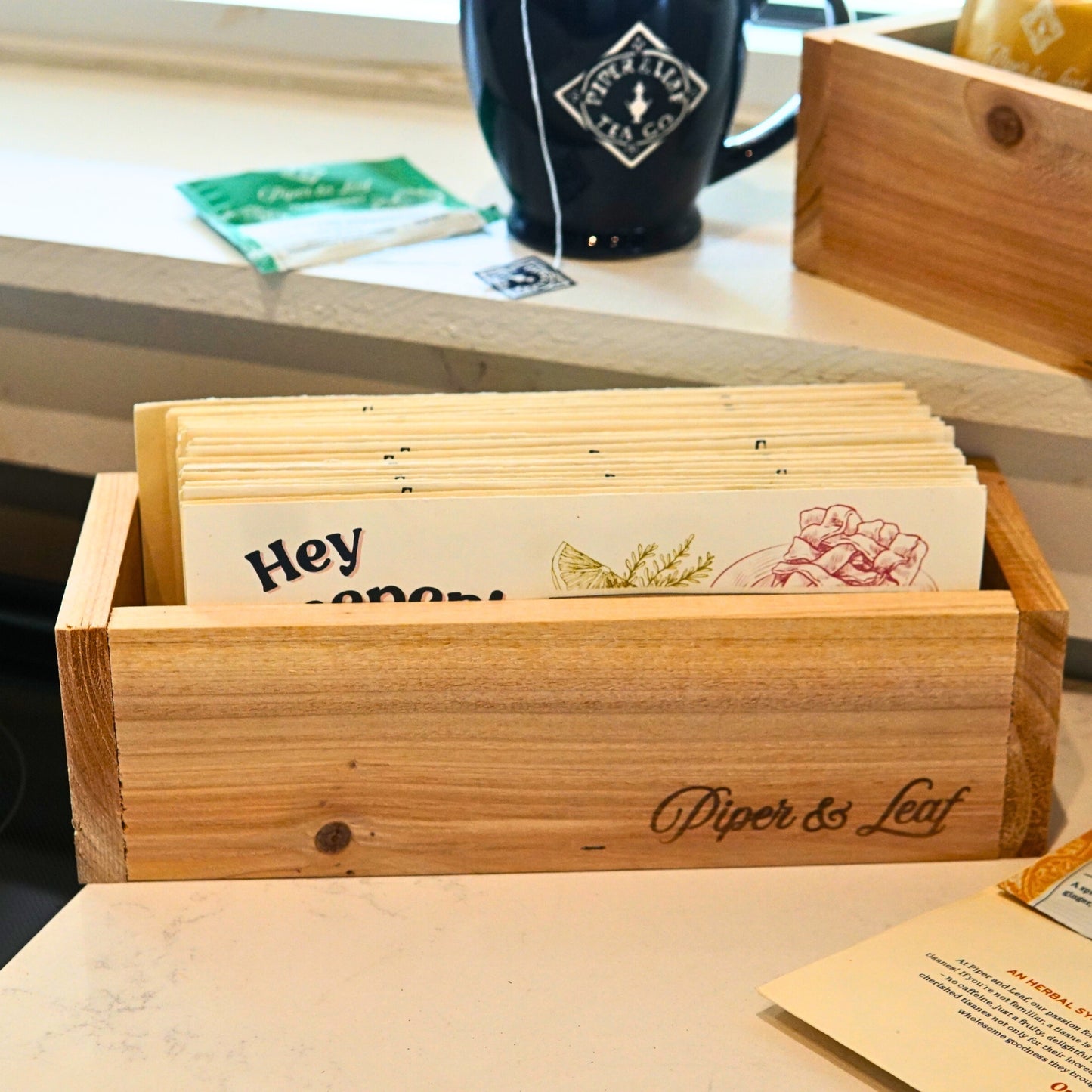 The "Wooden Steep Box" by Piper & Leaf Tea Co. neatly organizes tea bags beside a branded black mug. A green tea sachet sits nearby, ideal for your "3 Steeps a Week" routine.
