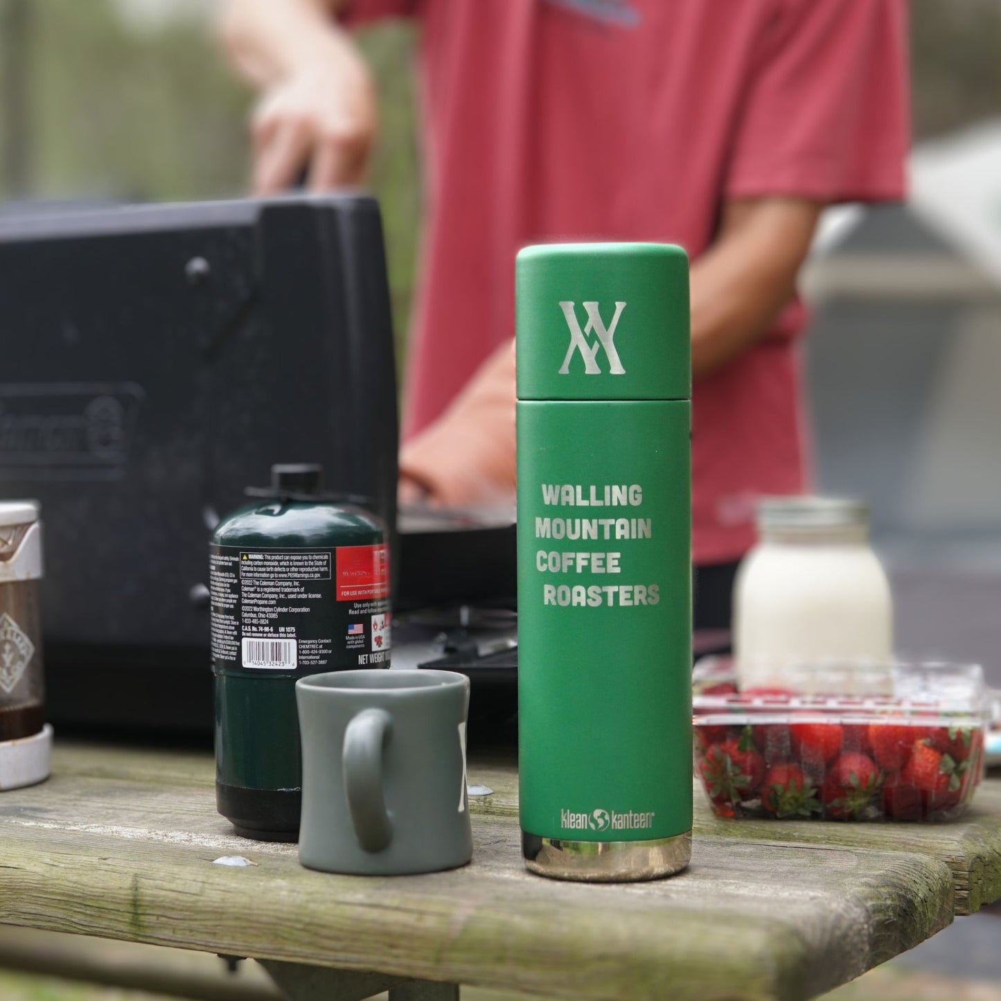 A WMF 32oz Thermos from Klean Kanteen, labeled "Walling Mtn. Coffee," rests on the picnic table next to a cup, strawberries, and a camping stove as someone expertly cooks in the background, embodying a true Klean Kanteen adventure.