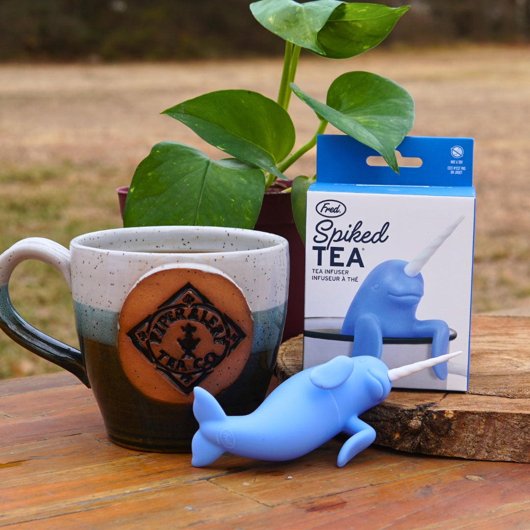 A charming Piper & Leaf Tea Co. Spiked Tea Infuser, shaped like a blue narwhal, sits beside a mug and its packaging on a wooden table with a plant providing an elegant backdrop.