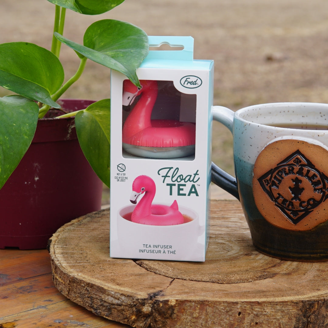 The Pool Flamingo Infuser by Piper and Leaf Tea Co. gracefully rests on a wooden surface, next to a "Float Tea" box, ready to steep your loose-leaf tea. It's the ideal gift for flamingo enthusiasts, sitting charmingly beside a mug and potted plant.