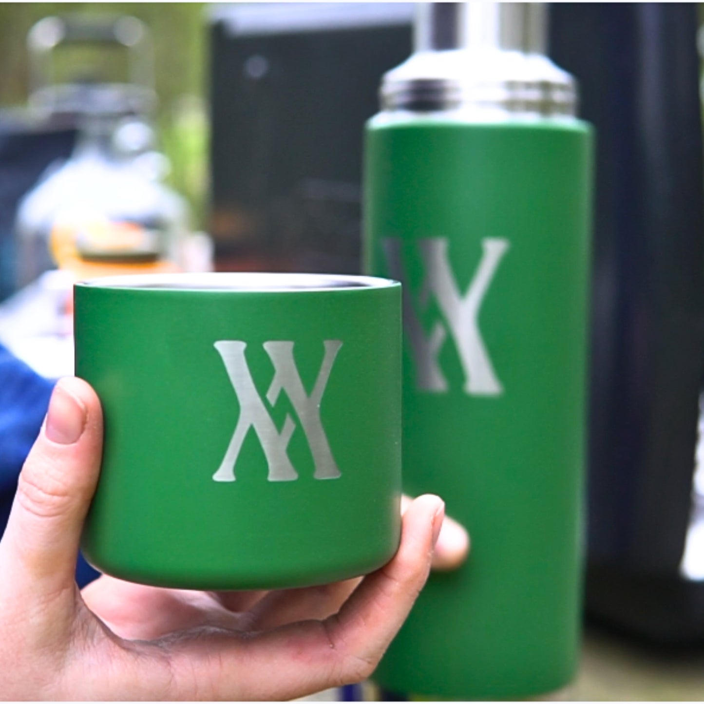 Someone holds a green cup with the Walling Mtn. Coffee logo next to a WMF 32oz Thermos from Klean Kanteen featuring the same design.