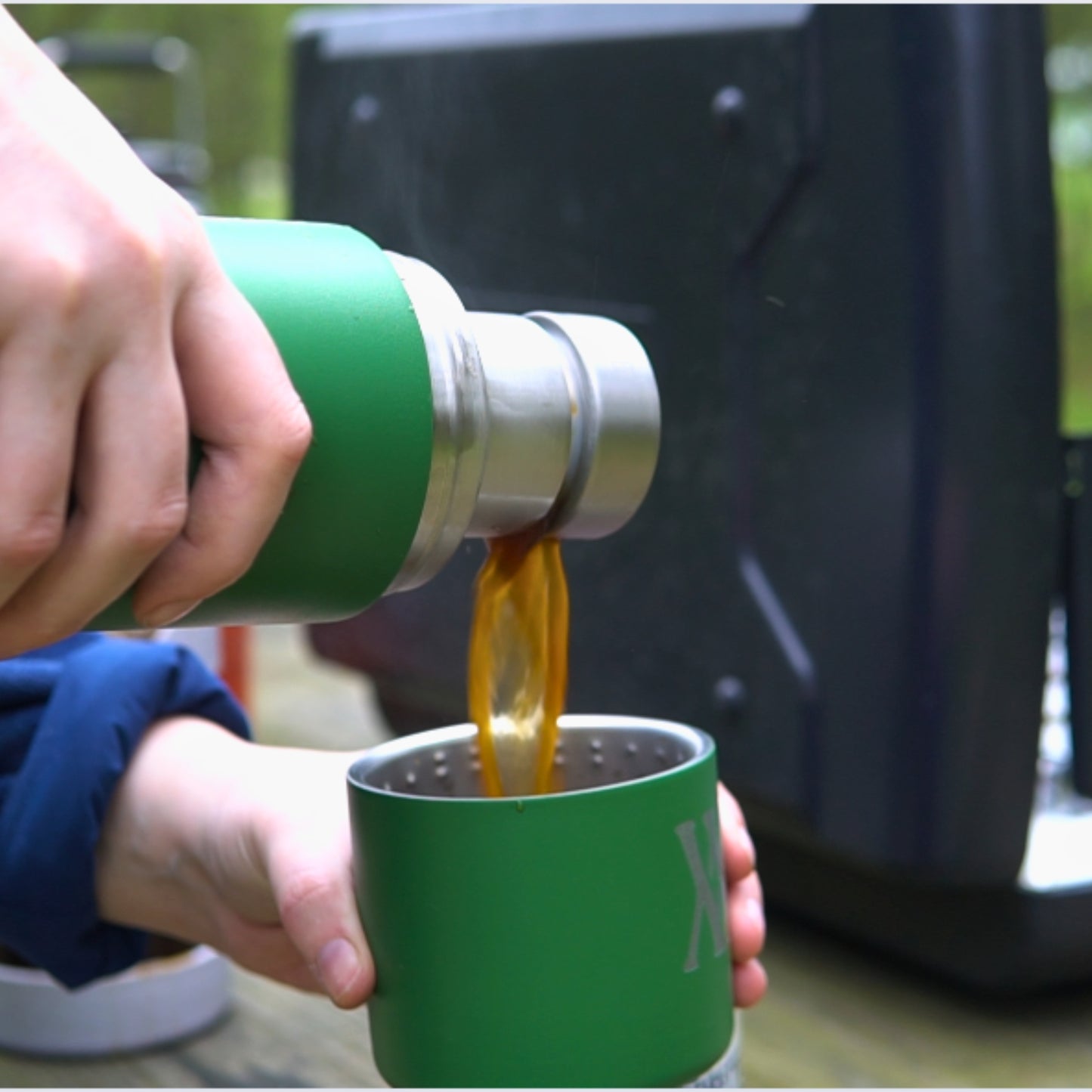 Outside, someone pours coffee from a green WMF 32oz Thermos by Klean Kanteen into a matching cup, while Walling Mtn. Coffee and camping gear are in the background.