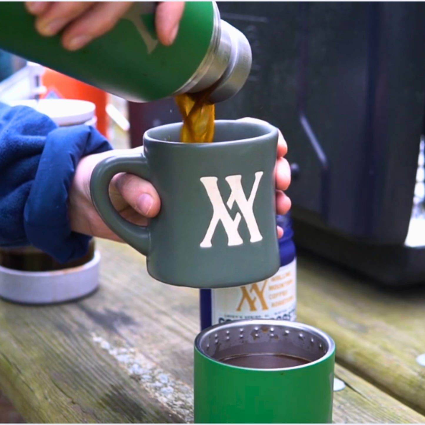 A person pours steaming coffee from a vibrant green WMF 32oz Thermos by Klean Kanteen into a Walling Mtn. Coffee logo mug, resting on an outdoor wooden table.