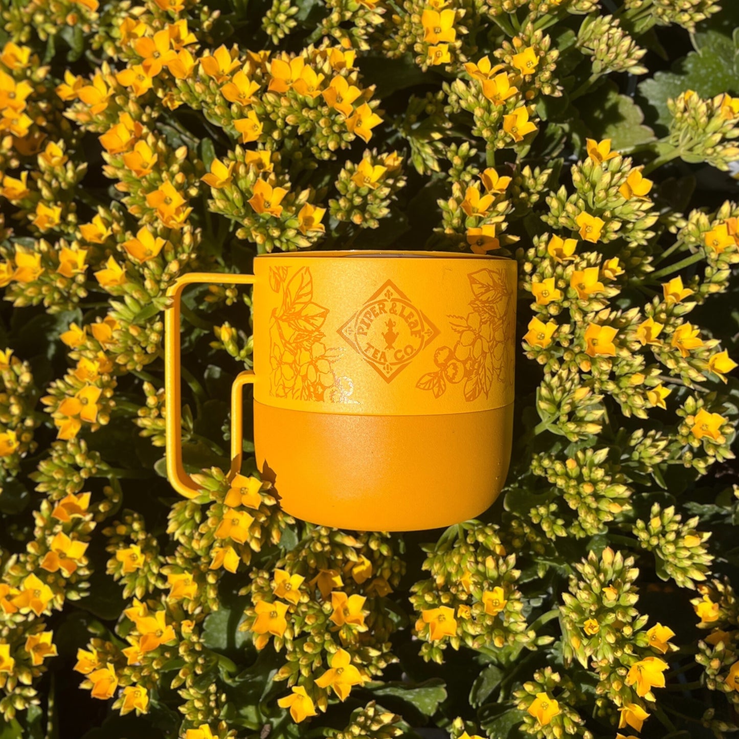 The P&L 14oz Rise Mug by Klean Kanteen from Piper & Leaf Tea Co., adorned with a floral logo, is surrounded by delicate yellow flowers and green leaves, making it an ideal companion for tea.