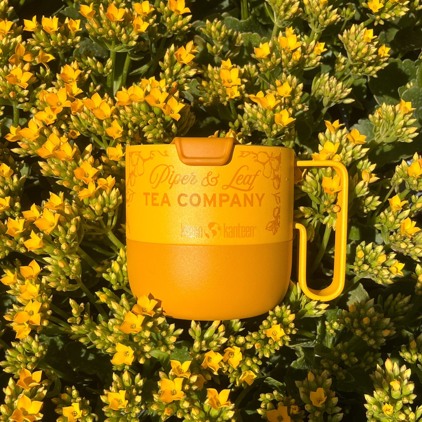 The P&L 14oz Rise Mug from Klean Kanteen by Piper & Leaf Tea Co., in vibrant yellow, sits amidst bright flowers in sunlight, making it the perfect companion for a relaxing afternoon.