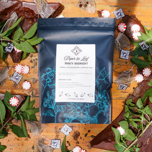 A 70ct bag of Piper & Leaf Tea Co.'s Minty Midnight Bulk Sachets is displayed among tea bags, mint leaves, chocolate pieces, and peppermint candies—ideal for a refreshing peppermint tea or soothing mint chocolate blend.