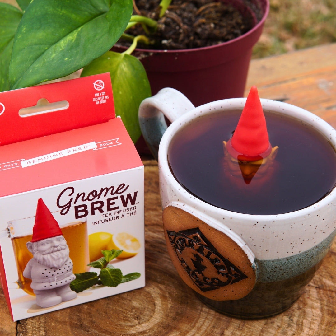 The Piper & Leaf Tea Co. Gnome Tea Infuser floats in a mug of tea next to its packaging on a wooden surface, with a potted plant in the background.