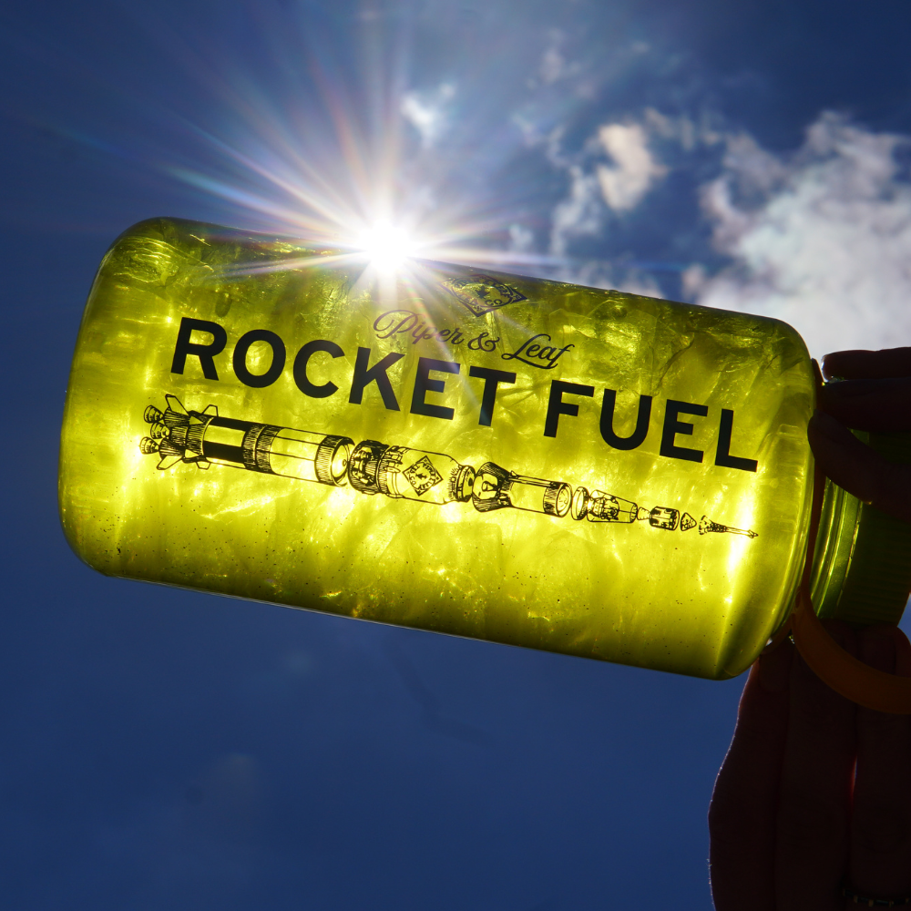 A Piper & Leaf Nalgene Water (Tea) Bottle labeled "Rocket Fuel Tea" is held up against the sun, creating a backlit effect.