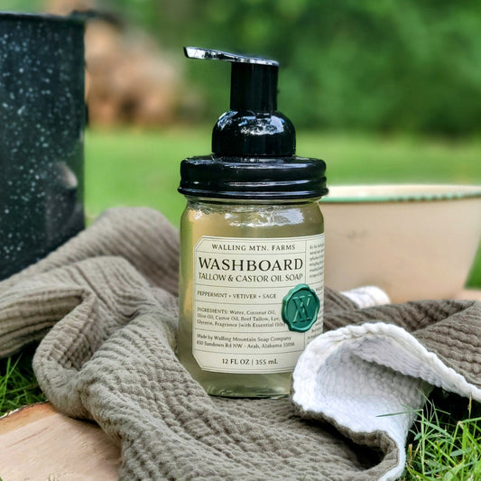 A 12oz pump bottle of Walling Mountain Farms Washboard Tallow & Castor Oil Foaming Hand Soap with peppermint, vetiver, and sage sits on a textured cloth outdoors, with a bowl and pot in the background.