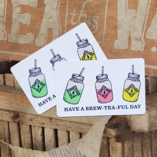 Three illustrated Piper and Leaf Tea Co. Physical P&L Gift Cards on a wooden surface featuring colorful drawings of mason jar drinks with the phrase "have a brew-tea-ful day.