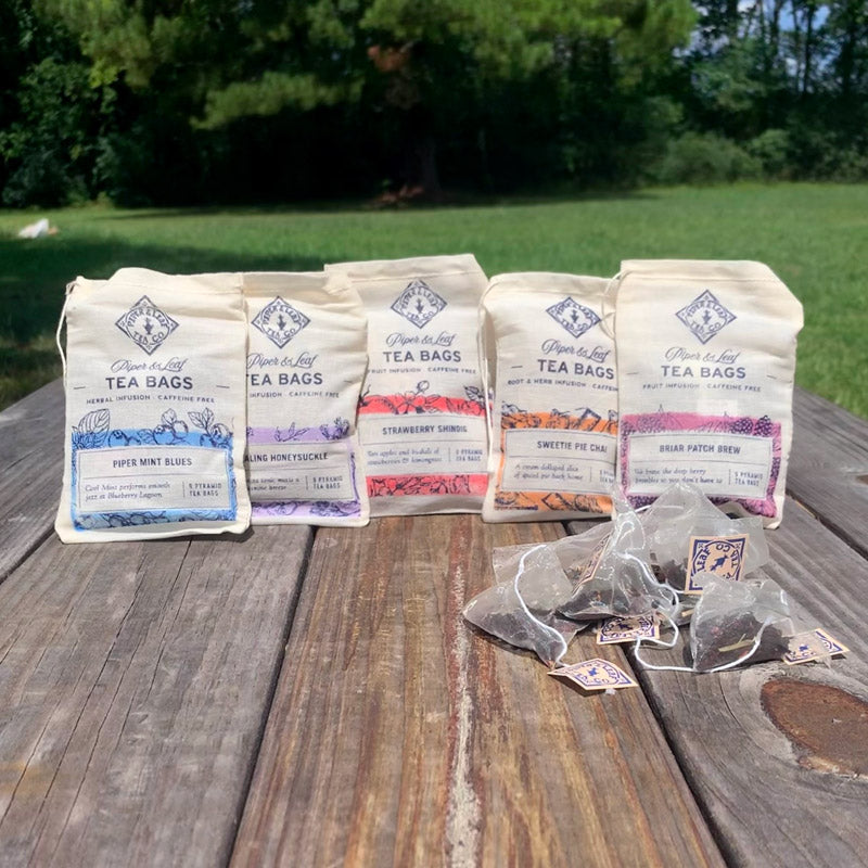 Five different Piper & Leaf muslin bags lined up on a table behind a small pile of tea bags