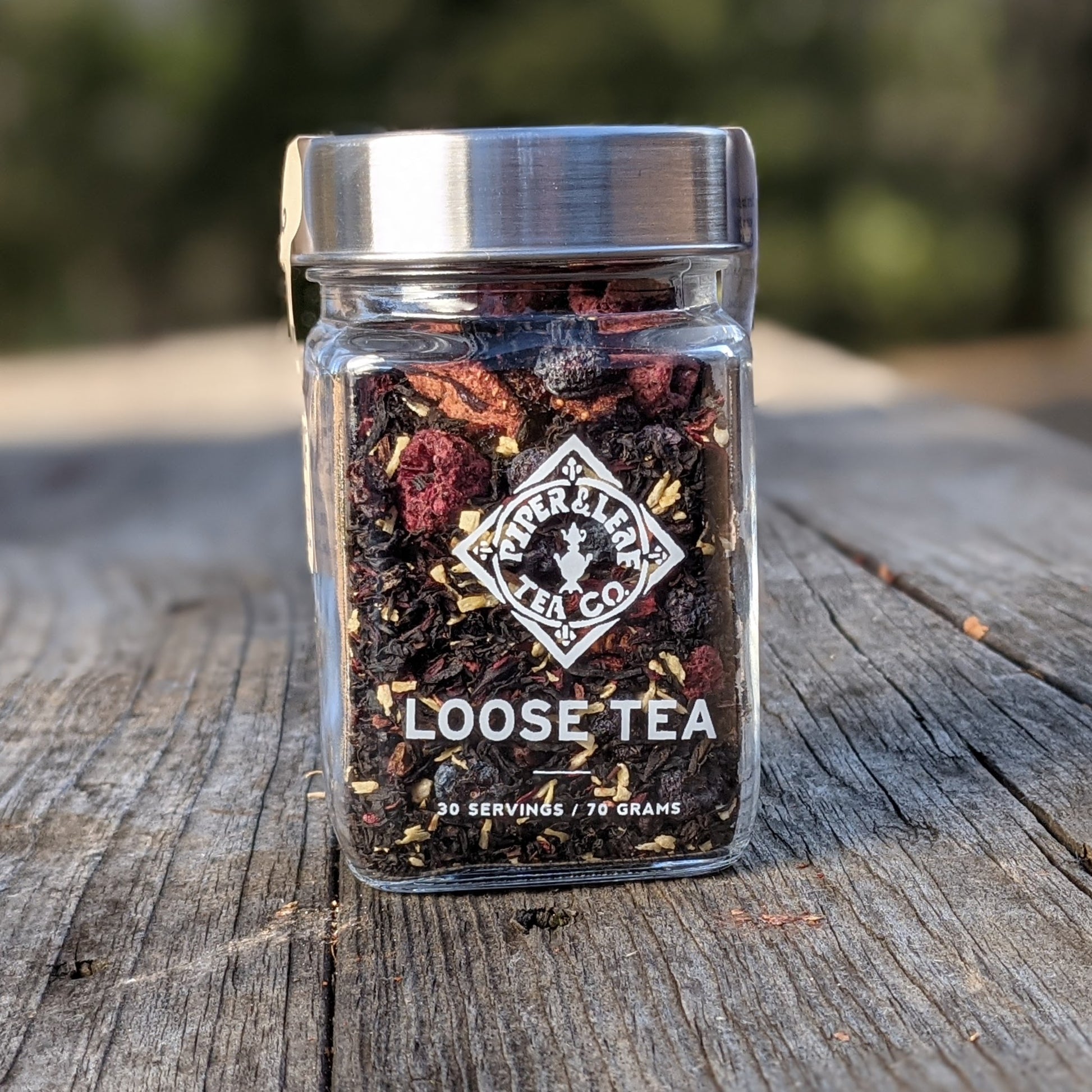 A glass jar labeled "Piper & Leaf Tea Co. Cherry Christmas Glass Jar of Loose Leaf Tea - 30 Servings" containing loose Ceylon Black tea leaves is placed on a wooden surface. The label notes "30 servings / 70 grams.
