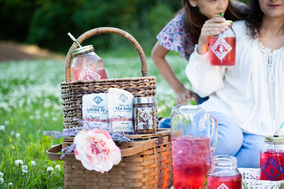 A woman and a girl drinking summer tea in a park, sitting beside a picnic basket filled with Piper & Leaf Tea Co. Sweet Dixie 9ct Tea Bags in Muslin and a glass jar of chamomile flowers.