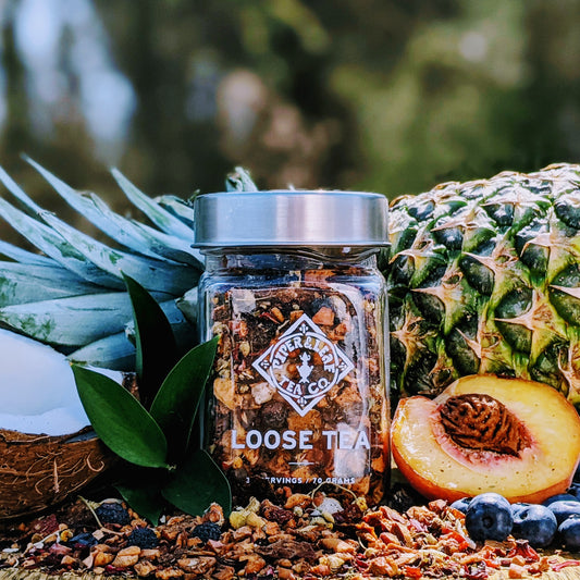 A Sweet Dixie Glass Jar of Loose Leaf Tea from Piper & Leaf Tea Co. sits among tropical fruits like pineapple, coconut, and peaches, surrounded by green leaves and blueberries.