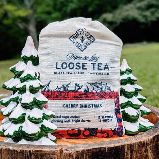 A "Piper & Leaf Tea Co." Cherry Christmas Muslin Bag of Loose Leaf Tea - 15 Servings is displayed outdoors surrounded by small decorative ceramic trees. The label notes it is a Ceylon Black tea blend with 15 servings per bag.