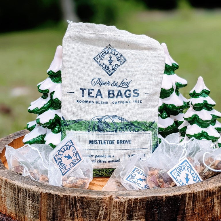 A bag of "Piper & Leaf Tea Co. Mistletoe Grove 9ct Tea Bags in Muslin" sits on a wooden surface, surrounded by small drawstring bags and decorative tree figurines. The label reads "Mistletoe Grove." This caffeine-free delight offers the rich experience of loose leaf tea in every sip.