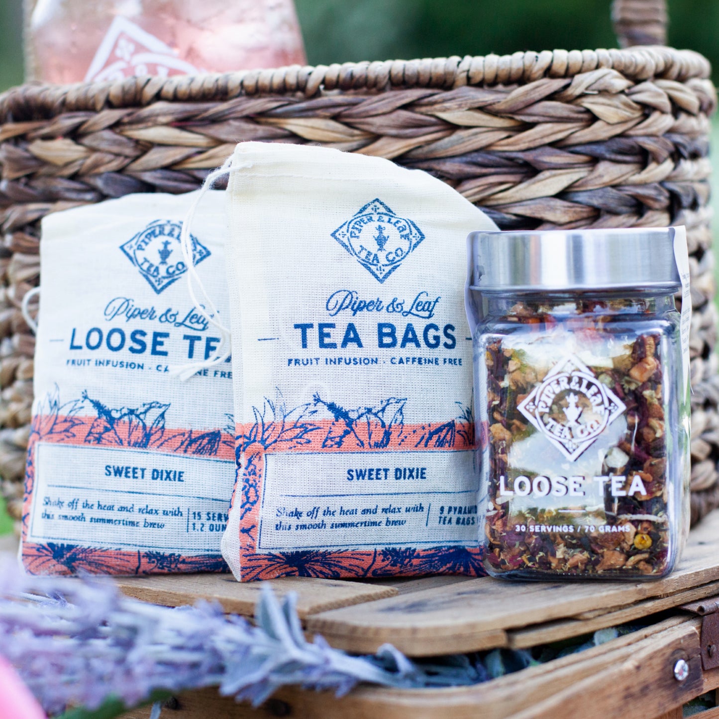 A wicker basket containing various labeled products including Sweet Dixie 9ct Tea Bags in Muslin and a jar of tea infusions from Piper & Leaf Tea Co., displayed outdoors on a wooden surface.