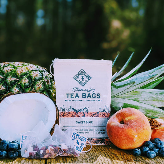 A display of Piper & Leaf Tea Co. Sweet Dixie 9ct Tea Bags in Muslin, fresh pineapple, peach, coconut half, and blueberries on a wooden surface with a forest backdrop.